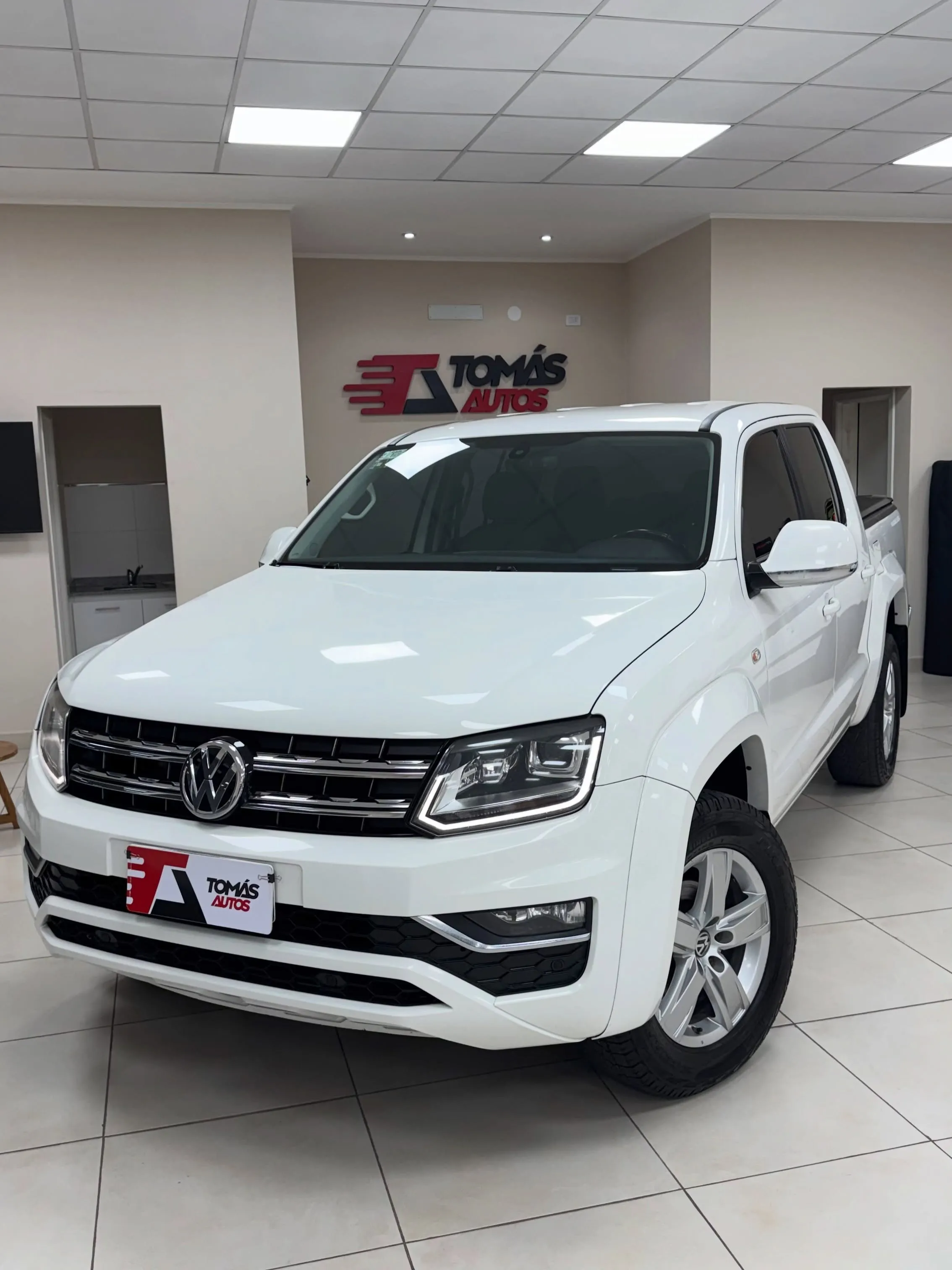 Volkswagen Amarok Comfortline 2018 blanca, de frente y perfil izquierdo, en el showroom de Tomás Autos